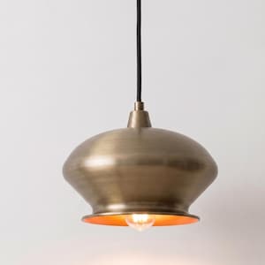 May include: A pendant light fixture featuring a brushed brass, dome-shaped metal shade with a wide, flared rim. The fixture is suspended by a black cord and includes a single, glowing light bulb visible within the open bottom of the shade.
