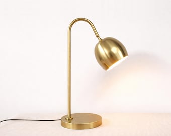 May include: A brass-colored metal table lamp featuring a slender, curved neck and a dome-shaped shade. The lamp has a circular base and emits a warm, bright light from the underside of the shade, which is directed downward at an angle.