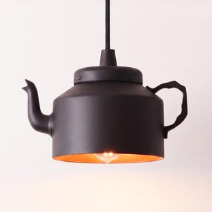 Op de afbeelding: Een matzwarte hanglamp in de vorm van een theepot, voorzien van een gebogen tuit, een getextureerd handvat en een cilindrisch lichaam. De binnenkant van de basis is afgewerkt in een warme kopertint, met een enkele lamp zichtbaar in het midden.