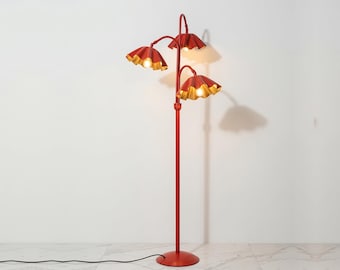 May include: A floor lamp with a tall, thin red metal pole and a circular base. It features three adjustable arms, each ending in a red, ruffled, flower-shaped shade with a metallic gold interior finish, housing a single glowing light bulb.