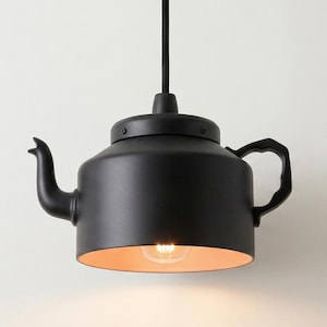 Könnte beinhalten: Eine mattschwarze Pendelleuchte in Form einer Teekanne. Das Äußere ist einfarbig schwarz mit einem geschwungenen Ausguss und Griff. Das Innere des Bodens ist in einem warmen, hellen Orangeton gehalten und eine leuchtende Glühbirne ist sichtbar.
