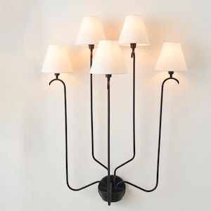 May include: A black metal wall sconce with five lights. Each light has a white lampshade. The sconce has a central base with curved arms extending to hold the lights. The design is minimalist and elegant, suitable for interior lighting.