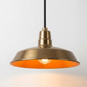 May include: A pendant light fixture featuring a wide, dome-shaped shade with a brushed brass finish. The interior of the shade is a warm, glowing orange hue, illuminated by a single exposed light bulb. A black cord extends from the top.