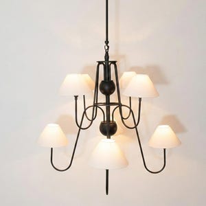 Puede incluir: Una lámpara de araña de metal negro con una varilla vertical central, dos detalles esféricos y seis brazos curvos que se extienden hacia afuera. Cada brazo sostiene una pantalla de tela blanca de forma cónica, creando una luminaria simétrica.