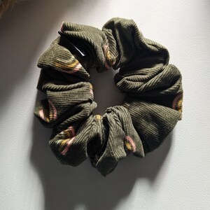May include: Olive green corduroy scrunchie with a repeating pattern of pink and yellow arches. The hair accessory is circular and gathered, with a black elastic band visible inside. A stylish hair tie for everyday wear.