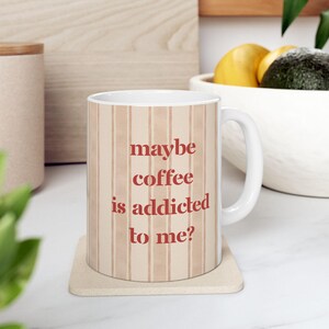 Könnte beinhalten: Ein weißer Keramikbecher mit einem vertikalen beigen Streifenmuster und dem roten Text "maybe coffee is addicted to me?" auf der Vorderseite. Der Becher hat einen Standardhenkel und steht auf einem quadratischen Untersetzer mit schlichtem Design.