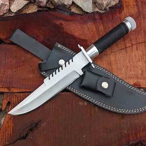 May include: A large, silver-bladed knife with a black handle and a black leather sheath. The knife has a serrated edge and a silver guard. The sheath has a small pouch and a belt loop. The knife is resting on a wooden surface.