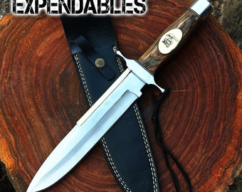 May include: A silver and brown hunting knife with a leather sheath. The knife has a wooden handle with a decorative design and a brass accent. The sheath has a stitched design and a strap. The knife is on a wooden surface with the text "THE EXPENDABLES" above it.