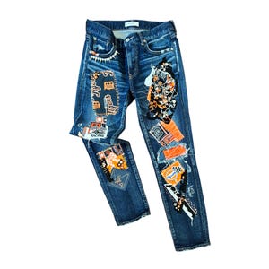 May include: Dark blue denim jeans featuring a variety of colorful patches and embroidered designs, including text such as Tigers, Racing, and Gulf. The pants have a distressed finish with frayed edges and a classic five-pocket construction.