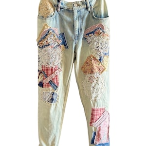 May include: Light-wash denim jeans with decorative patches. Patches include floral fabrics, lace doilies, and plaid patterns. The jeans have a relaxed fit and cuffed hems, showcasing a unique, handcrafted style.