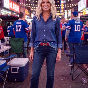 May include: A long-sleeve denim button-down shirt with a frayed hem, dark blue denim jeans, a red belt with a silver H-shaped buckle, and red leather loafers. The shirt features a blue and white trim along the bottom edge with the word BILLS.