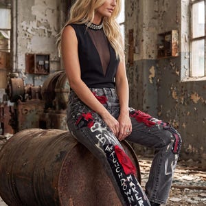 May include: A black sleeveless top with a sheer mesh panel and a jeweled neckline paired with grey denim jeans featuring red fabric patches, white graffiti-style text including "TOUCHDOWN" and "BAKER", and black lace-up boots.