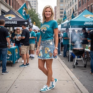 May include: A teal short-sleeved t-shirt featuring a Jacksonville Jaguars logo on the chest, paired with a light-wash denim mini skirt adorned with various fabric patches and a frayed hem, and white sneakers with teal accents on the soles and sides.