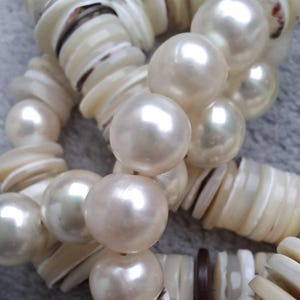 Könnte beinhalten: Nahaufnahme einer Halskette mit großen, glänzenden, cremefarbenen Perlen und muschelförmigen Perlen. Die Halskette hat eine natürliche, organische Ästhetik mit einer Mischung aus Texturen und Formen. Die Perlen haben einen weichen, reflektierenden Glanz.