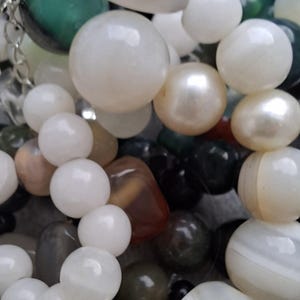 Puede incluir: Un surtido de cuentas de piedras preciosas de varias formas y tamaños. Las cuentas son principalmente blancas, verdes y negras, algunas con un brillo nacarado. Otros colores incluyen marrón y gris. Se ve una cadena plateada.