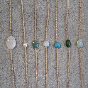 Könnte beinhalten: Sieben Edelsteinanhänger an beigefarbenen Kordeln. Die Edelsteine haben verschiedene Formen und Farben, darunter Weiß, Perle, Grün, Blau und Türkis. Die Anhänger sind vertikal vor einem grauen Hintergrund angeordnet.