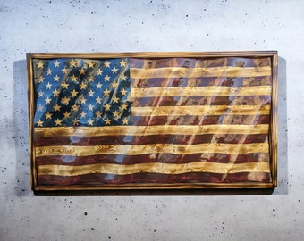 May include: A wooden American flag with red, white, and blue paint. The flag is framed in a dark wood frame and has a distressed look.