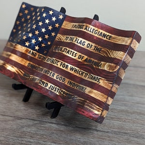 May include: A wooden American flag with the Pledge of Allegiance printed on it. The flag is displayed on a small black easel.