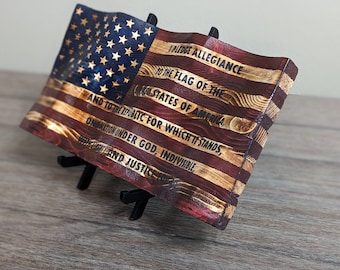 May include: Wooden American flag with the Pledge of Allegiance engraved on it. The flag features the stars and stripes design, with a blue field of stars and red and white stripes. The flag is displayed on a black stand.