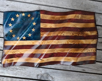 May include: A wavy wooden wall art piece shaped like a flag with a blue canton featuring thirteen stars arranged in a circle. The flag has alternating horizontal stripes in dark red and natural wood grain, with a glossy finish and rippled texture.