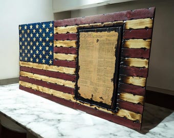 Op de afbeelding: Een Amerikaanse houten vlag met een verweerde afwerking. De vlag is gemaakt van afzonderlijke houten planken en heeft een rood, wit en blauw kleurenschema. De vlag is aan een muur gemonteerd en heeft een ingelijst exemplaar van de Onafhankelijkheidsverklaring in het midden.