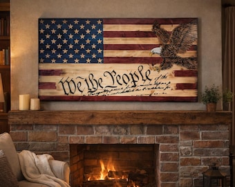 May include: Wooden American flag wall art with a distressed finish. The flag features the stars and stripes, an eagle, and the words "We the People" from the Constitution. The artwork is displayed above a brick fireplace.
