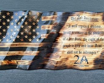 Pode incluir: Uma peça de arte de parede em madeira em forma de bandeira americana ondulada com acabamento queimado. O lado esquerdo apresenta estrelas e listras, enquanto o lado direito exibe o texto do 2nd Amendment e o símbolo 2A 1791 em letras pretas.