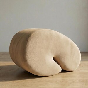 Op de afbeelding: Een beige, niervormige poef met een afgeronde vorm en een gebogen inkeping. Het zachte, gestoffeerde stuk staat op een houten vloer en biedt een minimalistisch design. De neutrale kleur en unieke vorm maken het tot een veelzijdig meubelstuk.