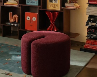 Könnte beinhalten: Ein gemütliches Wohnzimmer mit einem türkisfarbenen Teppich, einem weinroten Hocker und einem dunklen Holzregal voller Bücher und Dekorationsgegenstände. Kunstwerke schmücken die Wände, und ein blauer Sessel steht in der Ecke. Eine moderne Lampe hängt von der Decke.