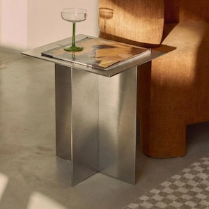 Op de afbeelding: Een moderne bijzettafel van geborsteld metaal met een vierkant blad. Een helder coupeglas met een groene steel staat op de tafel, die ook een tijdschrift bevat. Op de achtergrond een oranje fauteuil.