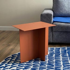 May include: A modern, terracotta-colored side table with a rectangular top and a single, wide leg. The table is made of a solid material and stands on a blue and white patterned rug. The table's height is approximately 24 inches.