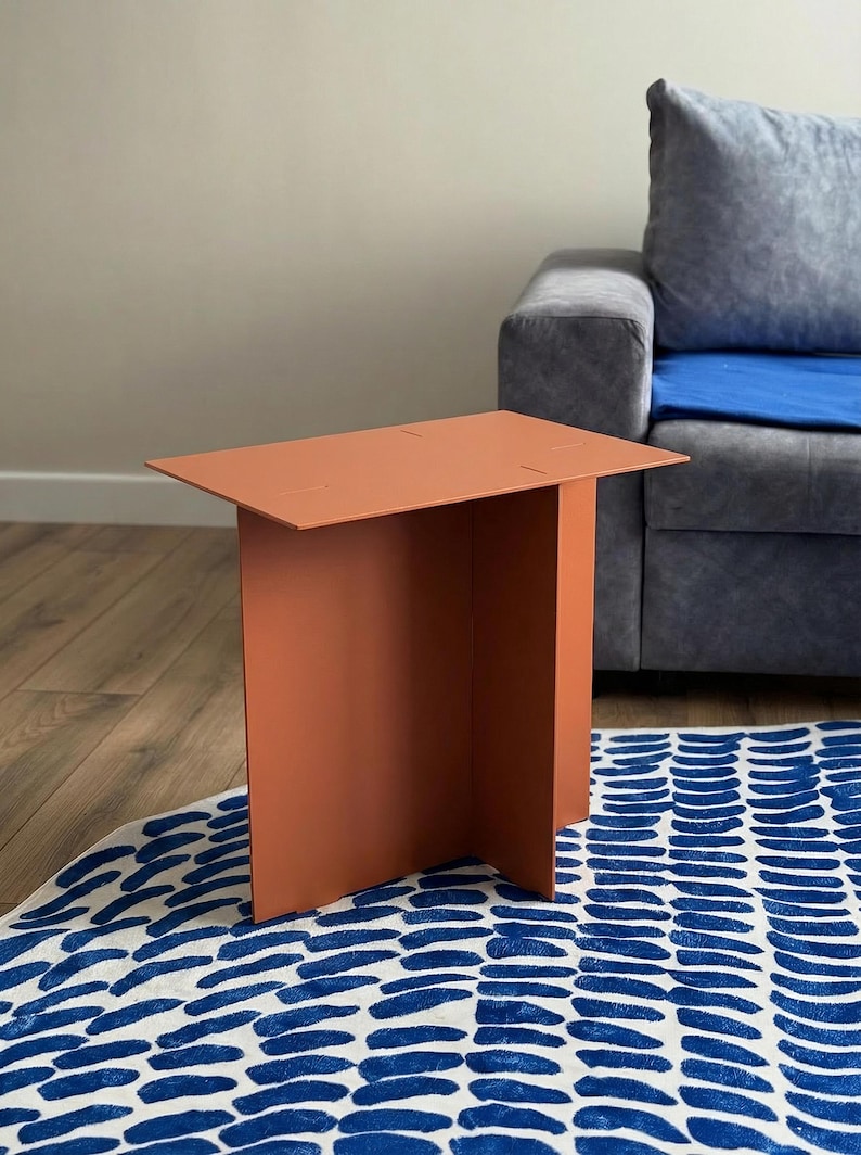 May include: A modern, terracotta-colored side table with a rectangular top and a single, wide leg. The table is made of a solid material and stands on a blue and white patterned rug. The table's height is approximately 24 inches.