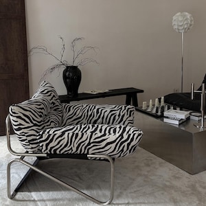 Puede incluir: Un sillón con tapicería de tela con estampado de cebra en blanco y negro. La silla está sostenida por una estructura de metal cromado pulido con un diseño de base geométrico distintivo, que ofrece un aspecto estructurado con asientos acolchados.