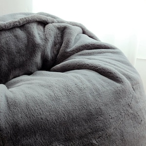 May include: A large, gray beanbag chair with a soft, faux fur texture. The plush material is a solid, medium gray color, with subtle variations in tone. The beanbag is round and appears comfortable.