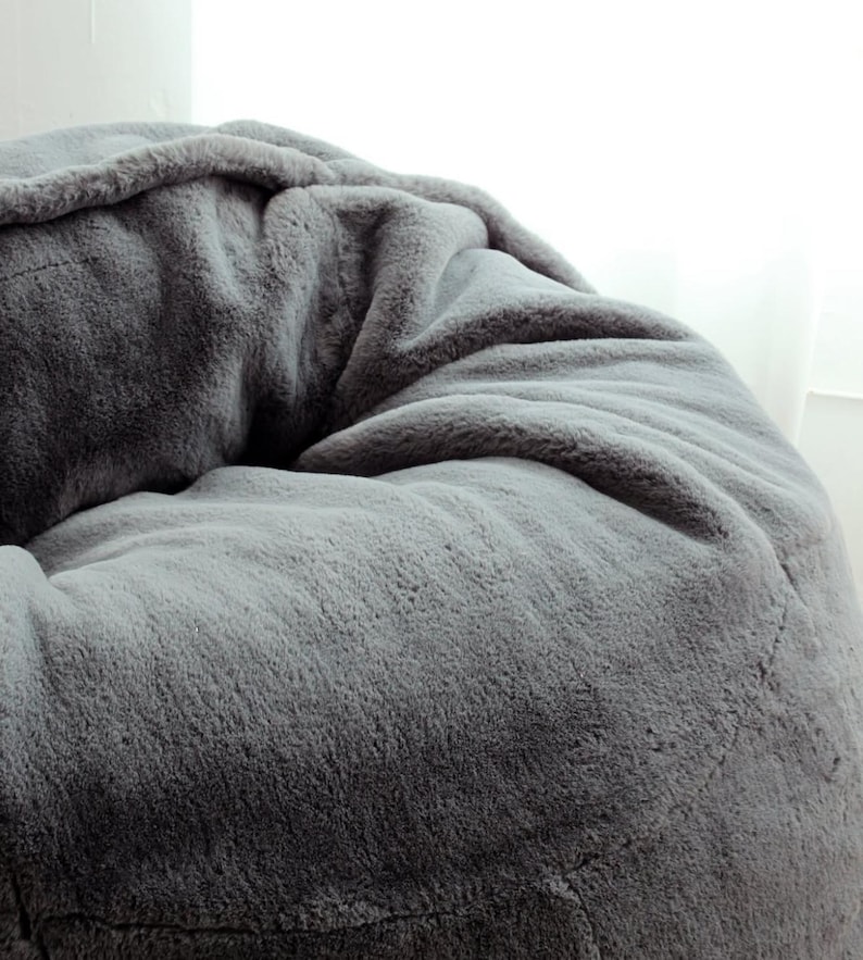 May include: A large, gray beanbag chair with a soft, faux fur texture. The plush material is a solid, medium gray color, with subtle variations in tone. The beanbag is round and appears comfortable.