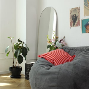 May include: A gray beanbag chair with a red and white striped pillow. A tall, arched mirror leans against the white wall. A potted plant and a small black marble side table are also in the room. Artwork hangs on the wall.
