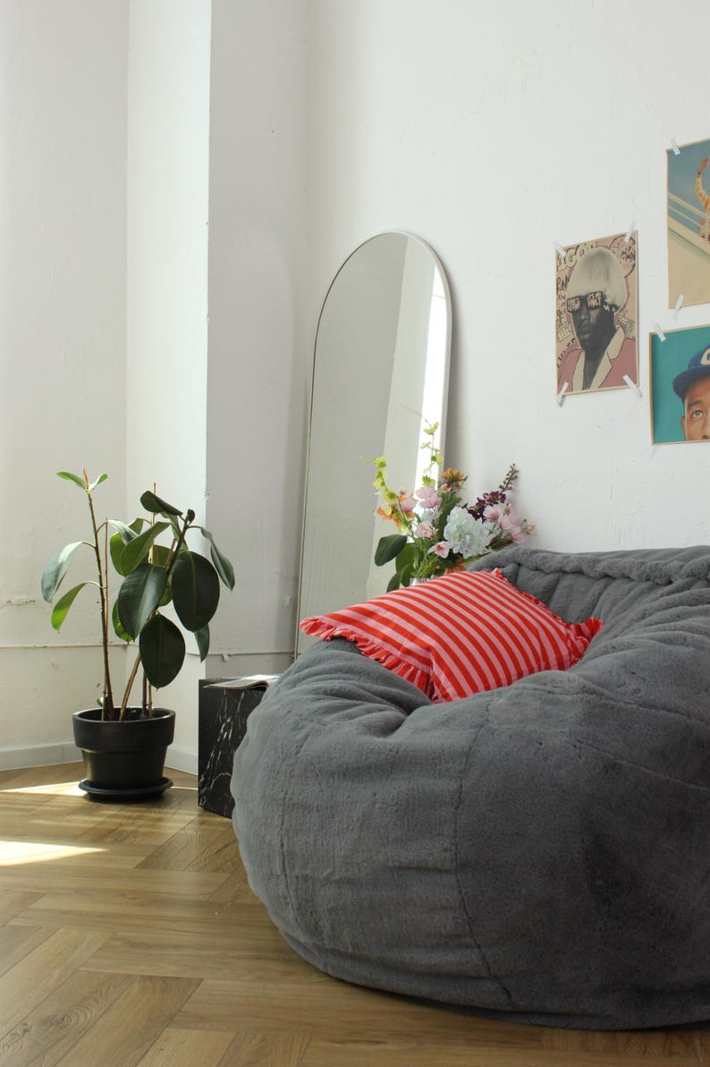May include: A gray beanbag chair with a red and white striped pillow. A tall, arched mirror leans against the white wall. A potted plant and a small black marble side table are also in the room. Artwork hangs on the wall.