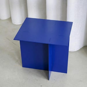 May include: A vibrant blue, geometric side table. The table is constructed from folded metal sheets, forming a boxy shape with a flat top. The design features subtle cut lines. The table is set against a neutral background.