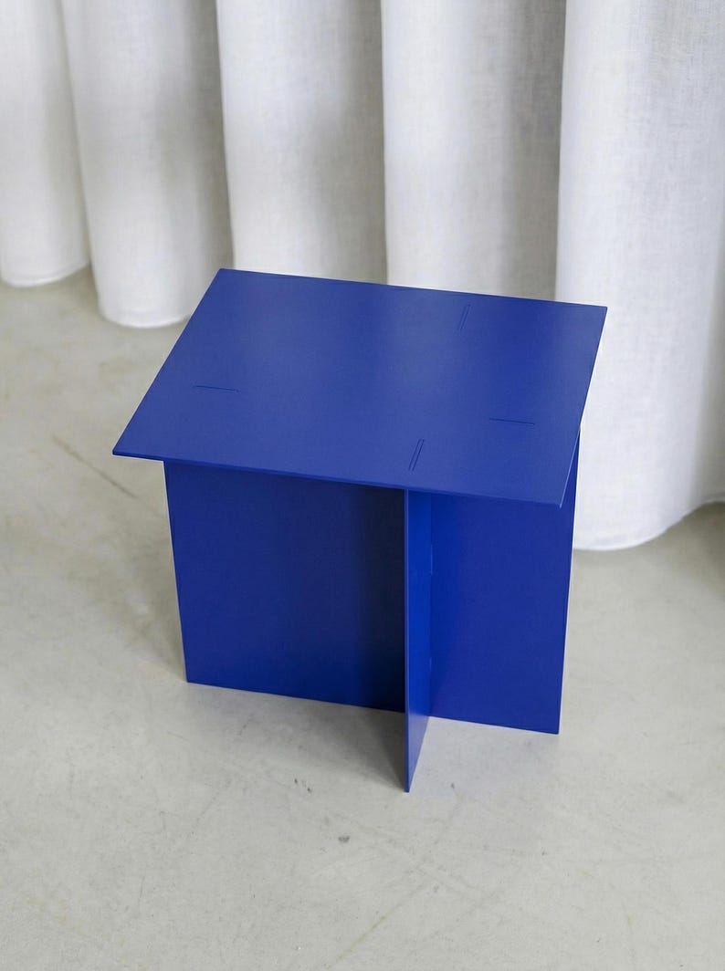 May include: A vibrant blue, geometric side table. The table is constructed from folded metal sheets, forming a boxy shape with a flat top. The design features subtle cut lines. The table is set against a neutral background.