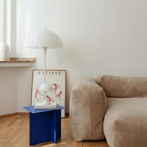 May include: A modern interior scene featuring a white mushroom-shaped floor lamp and a smaller table lamp. A vibrant blue side table holds books. A beige corduroy sofa and a framed "MATISSE" poster complete the room's decor.