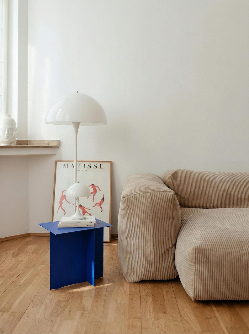 May include: A modern interior scene featuring a white mushroom-shaped floor lamp and a smaller table lamp. A vibrant blue side table holds books. A beige corduroy sofa and a framed "MATISSE" poster complete the room's decor.