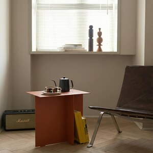 May include: A modern interior scene featuring a burnt orange side table with a black teapot and a cup and saucer. A black Marshall speaker sits on the floor. A brown leather chair and a window with blinds are in the background.