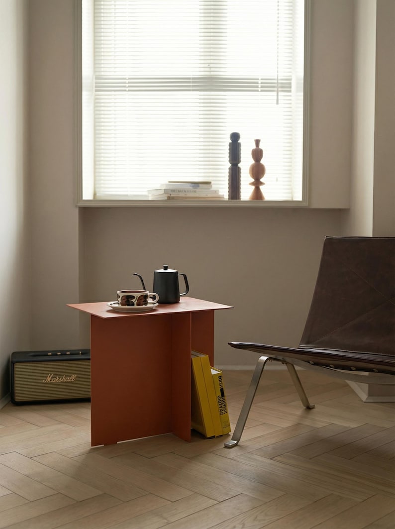 May include: A modern interior scene featuring a burnt orange side table with a black teapot and a cup and saucer. A black Marshall speaker sits on the floor. A brown leather chair and a window with blinds are in the background.