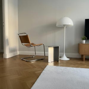 May include: A modern living room featuring a brown leather chair with a chrome frame, a small silver side table, and a white floor lamp. The room has light gray walls, a wooden floor, and a white rug.