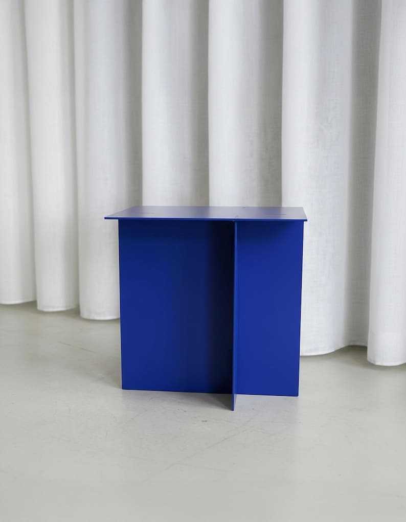 May include: A modern, cobalt blue side table with a minimalist design. The table has a rectangular top and a solid, geometric base. The table is made of a solid material and is suitable for various interior design styles.