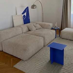 May include: A modern living room setup featuring a beige corduroy sectional sofa, a blue side table, and a matching ottoman. A blue abstract art piece leans against the wall. A silver arc lamp provides lighting.