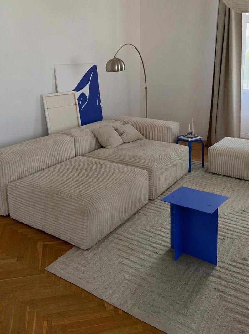 May include: A modern living room setup featuring a beige corduroy sectional sofa, a blue side table, and a matching ottoman. A blue abstract art piece leans against the wall. A silver arc lamp provides lighting.