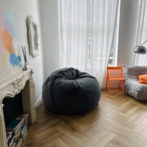 May include: A cozy living room scene featuring a large, dark gray beanbag chair. The room has a white fireplace, a stack of books, and a modern orange chair. The floor is a light brown herringbone pattern.