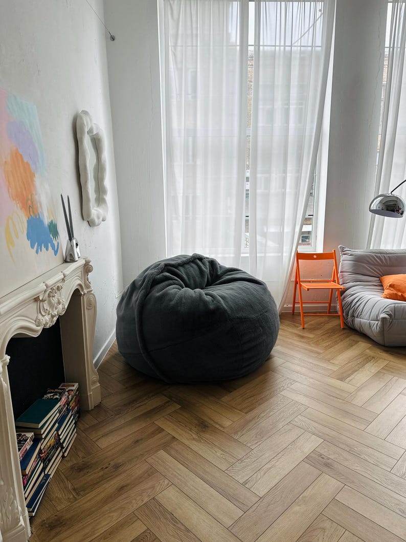 May include: A cozy living room scene featuring a large, dark gray beanbag chair. The room has a white fireplace, a stack of books, and a modern orange chair. The floor is a light brown herringbone pattern.
