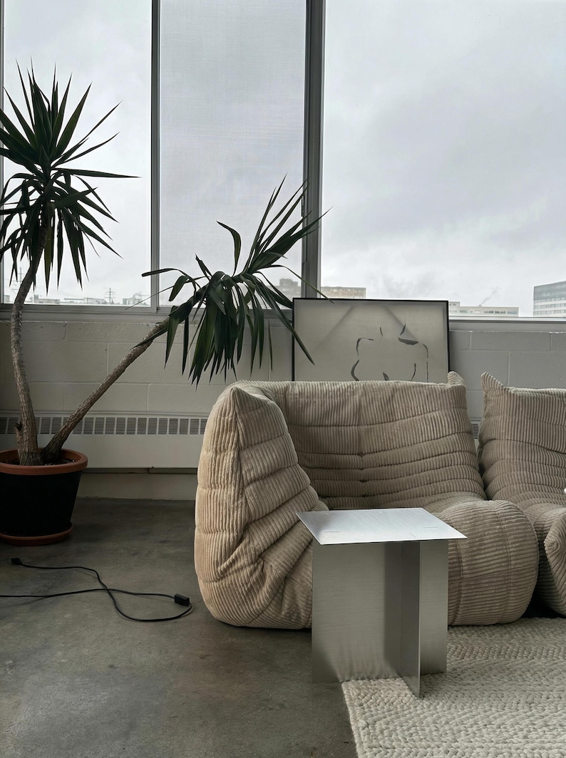 May include: A modern living room scene featuring a beige corduroy sofa, a small square metal side table, and a potted plant with long green leaves. A framed artwork and a window are in the background. The room has a minimalist aesthetic.